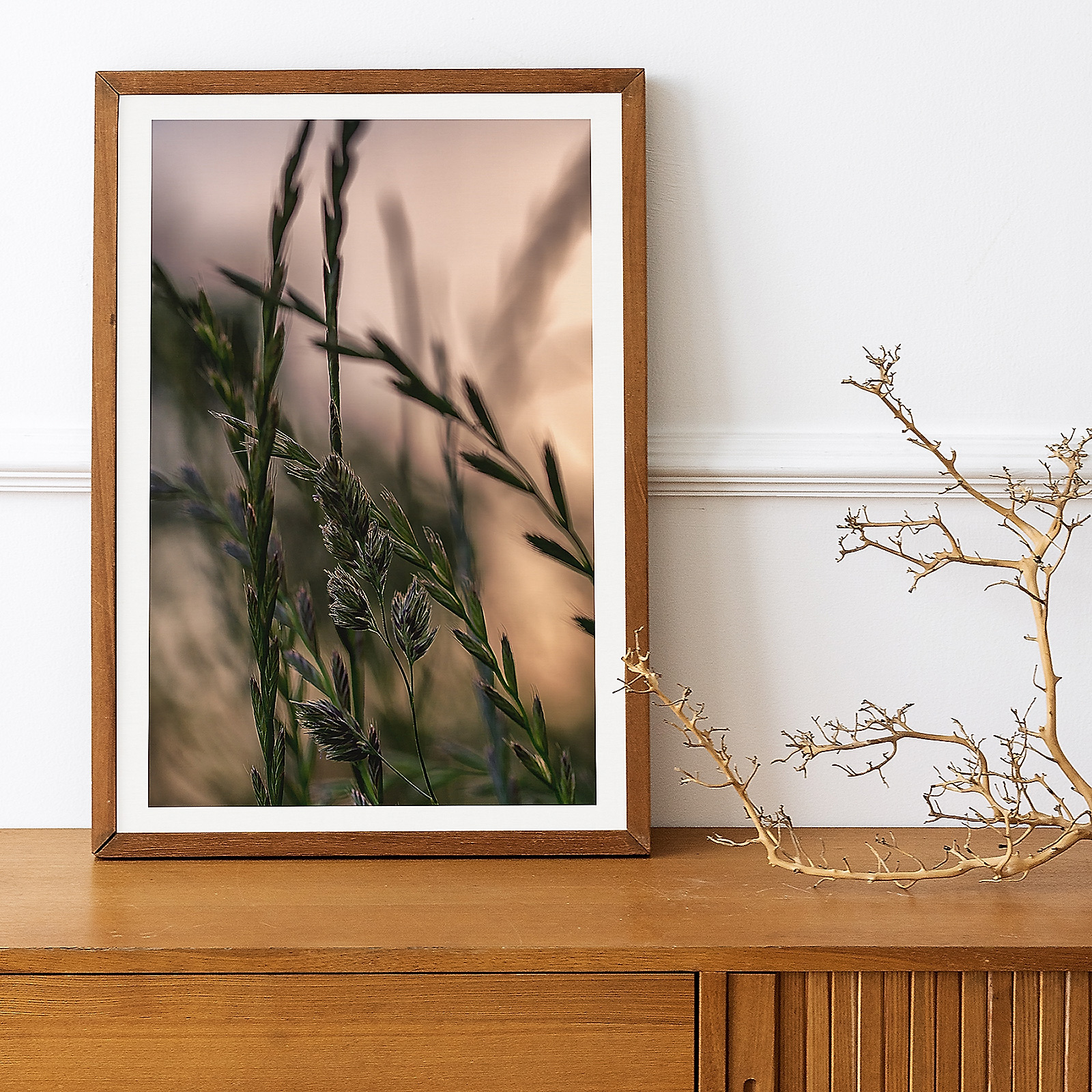 Close-up macro photograph of meadow grass in evening light, framed in a light wooden frame, standing on top of a wooden sideboard. A wooden bare branch decorates the right side of the sideboard.