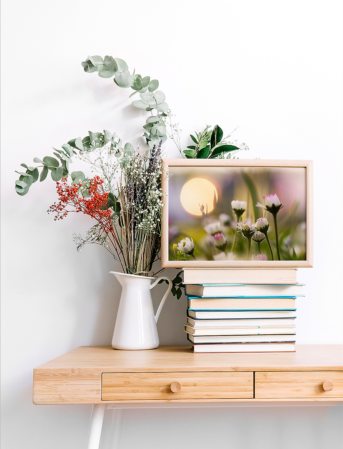 White-framed artwork 'Faintly Sweet' resting on a stack of books atop a modern wooden table, accompanied by a vase of wildflowers.