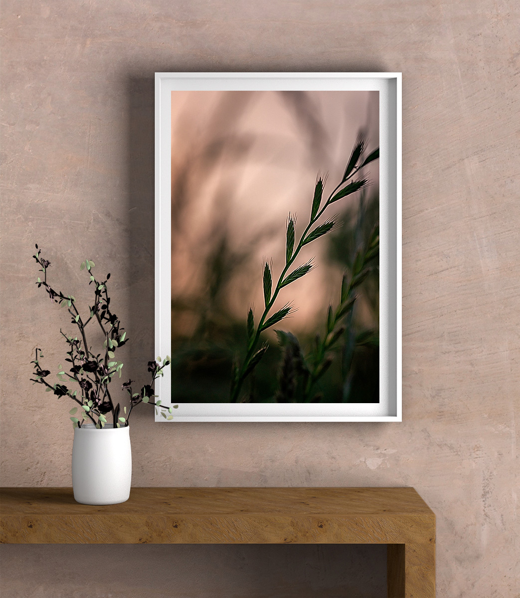 Close-up macro photograph of meadow grass in evening light, framed in a white frame with white mounting, hanging on a wall. A wooden sideboard and a leafy plant in a white pot are visible underneath.